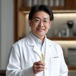 白衣を着た、経験豊かな中年の日本人男性化粧品職人が、ガラス容器に入った化粧液を慎重に調合しているクローズアップショット。彼の目には集中と情熱が宿っている